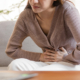 A woman sitting on the sofa gripping her abdomen in pain with both hands.